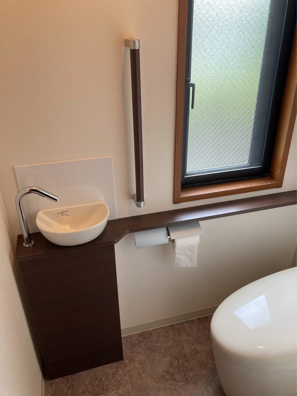 M様邸トイレ完成。手洗いキャビネットと紙巻き器の設置状況。ブラウン系の木目でまとめた上質な仕上がりです。