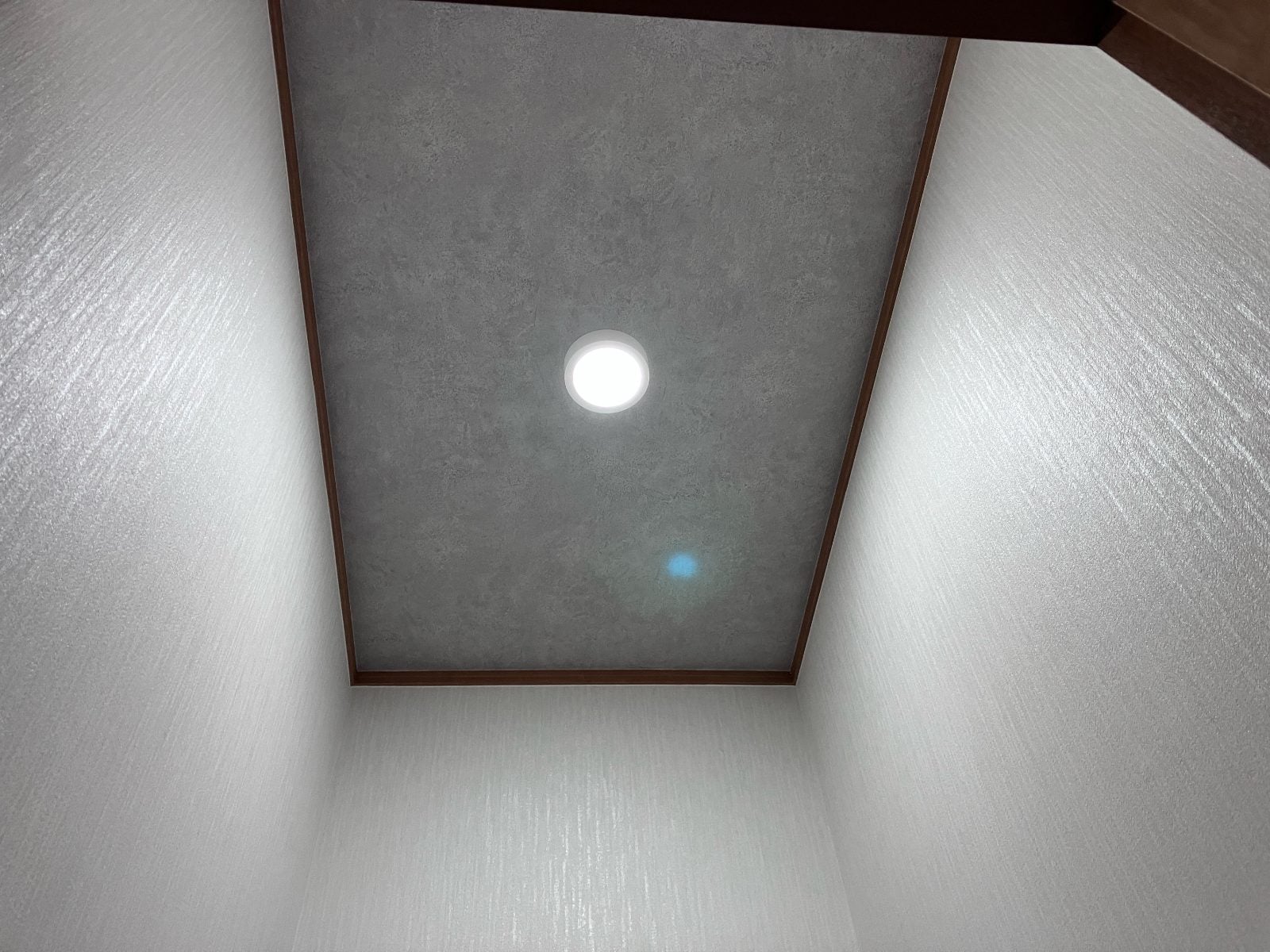 I様邸トイレ施工後の天井まわり。グレーのアクセントクロスとダウンライトでモダンな仕上がりになっています。